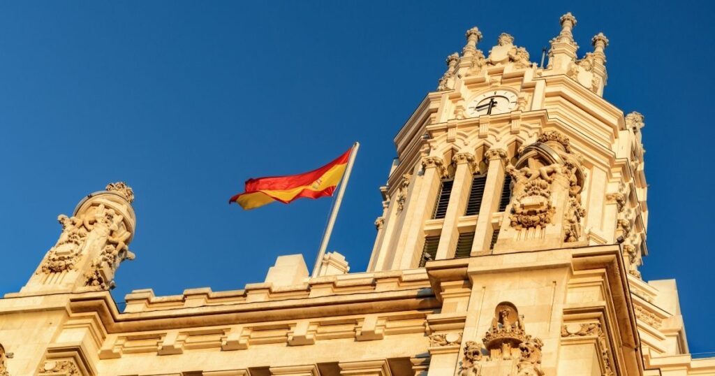 A wide 16:9 scenic view of a majestic historic building in Spain with the Spanish flag flying at the top under a clear blue sky.