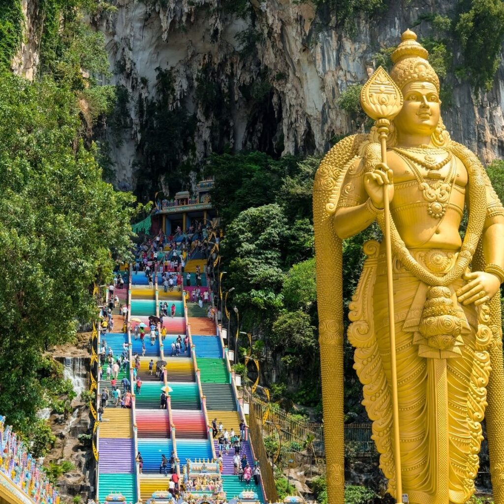 The dramatic entrance of the Batu Caves shrine in Kuala Lumpur, Malaysia, featuring the massive gold Lord Murugan statue standing in front of the iconic, vibrant rainbow-coloured staircase leading up into the limestone cavern.