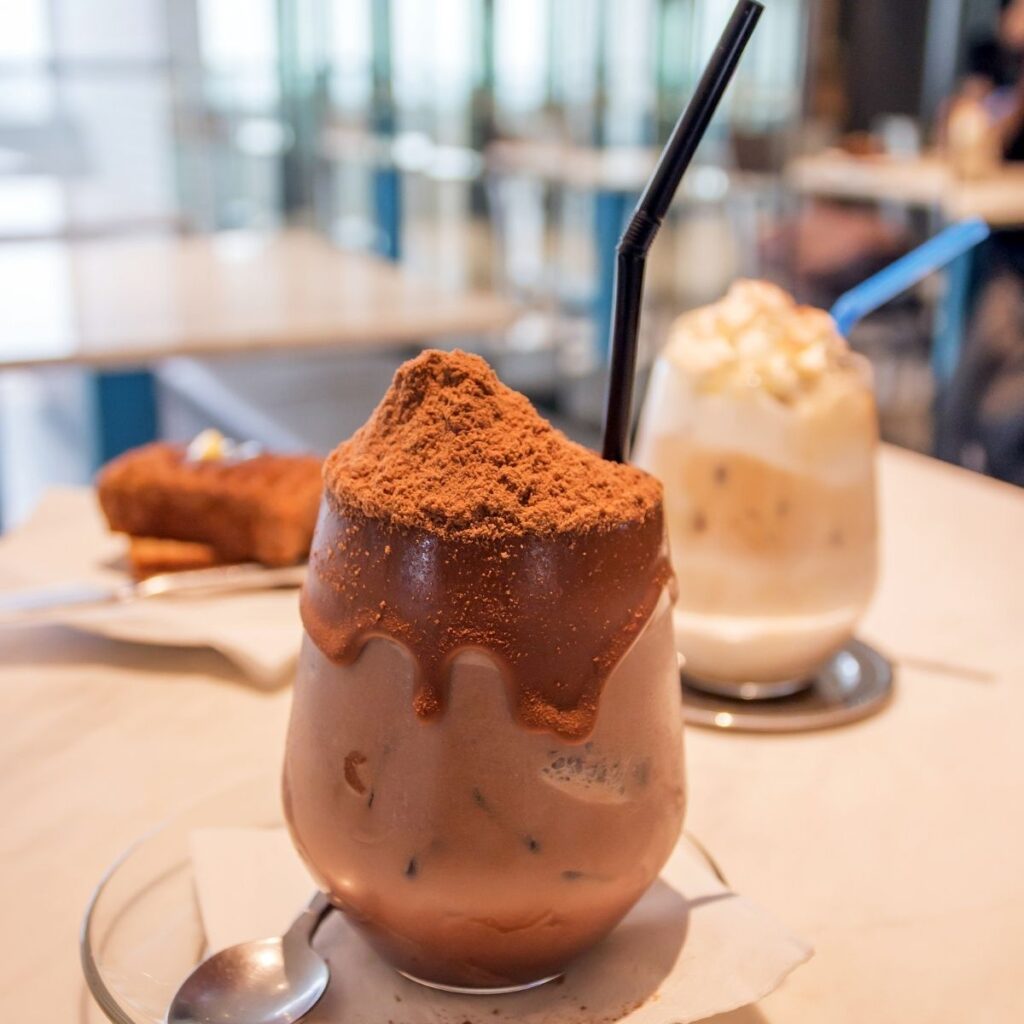 A close-up of a Milo Dinosaur drink in a small glass cup, showing the brown malty chocolate beverage layered under a mountain of dry Milo powder on top, with a bit of the chocolate drink lingering down the side of the glass