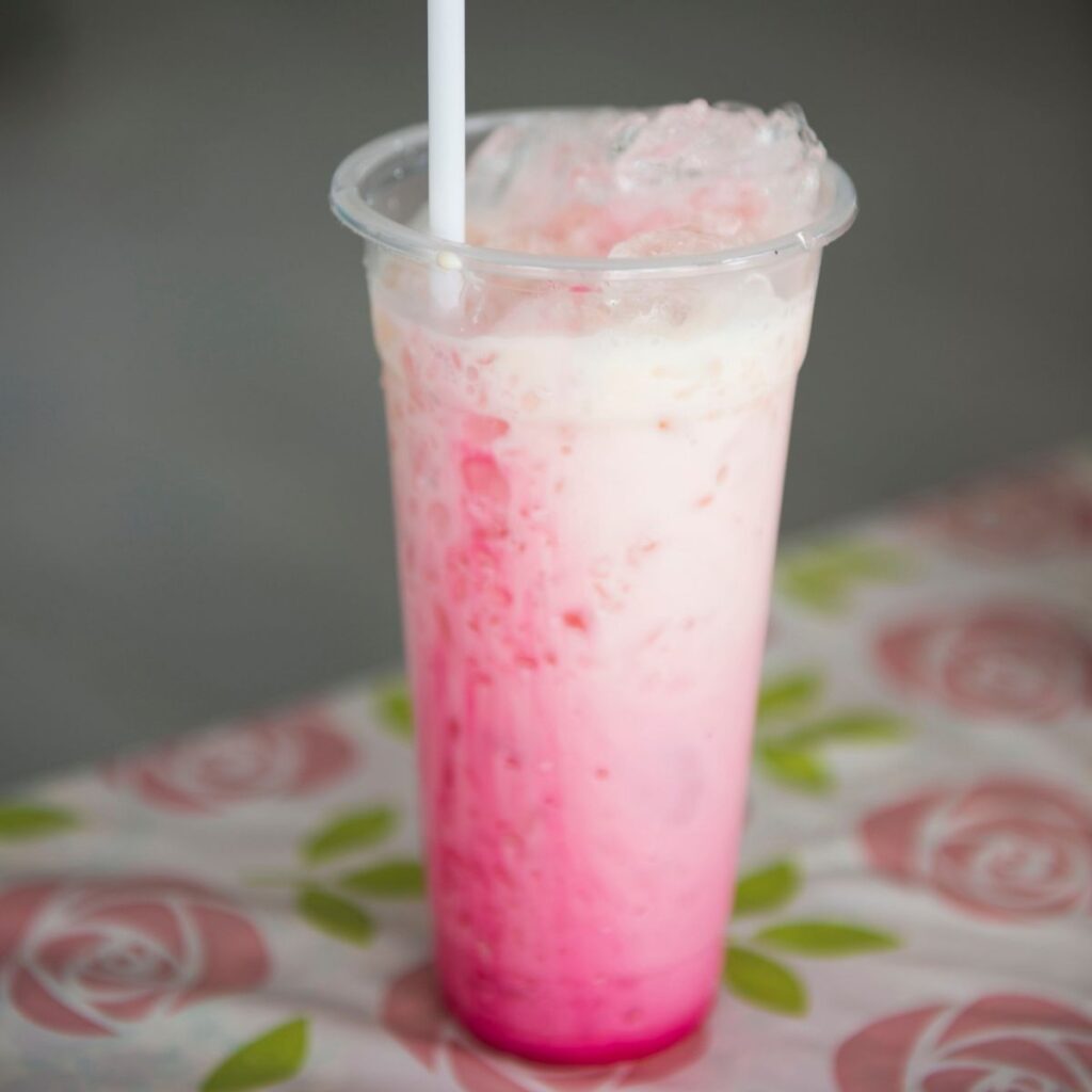 A vibrant Sirap Bandung drink showing a beautiful gradient of pink rose syrup mixing with creamy white milk inside a glass, resting on a decorative rose petal pattern tablecloth.