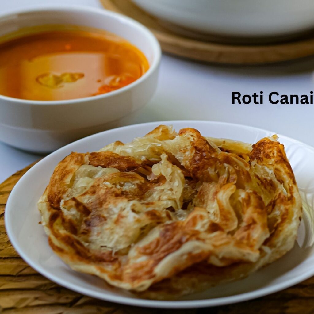 A plate of flaky, crispy Malaysian Roti Canai flatbread served alongside a side bowl of rich, aromatic orange-red curry sauce for dipping.