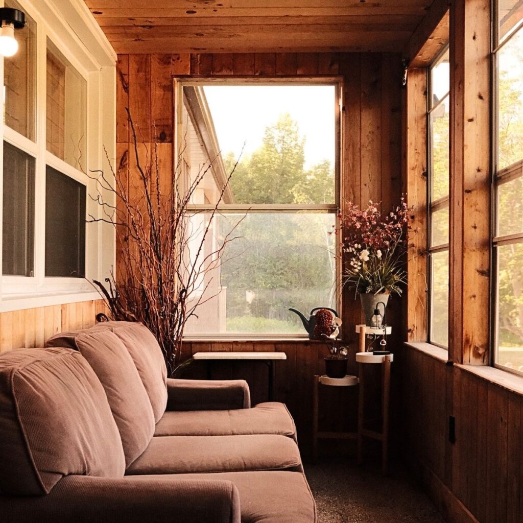 A cozy wood cottage vestibule in Innisfil, Ontario, featuring a warm brown couch and a scenic view of a golden lake sunset visible through the large window.
