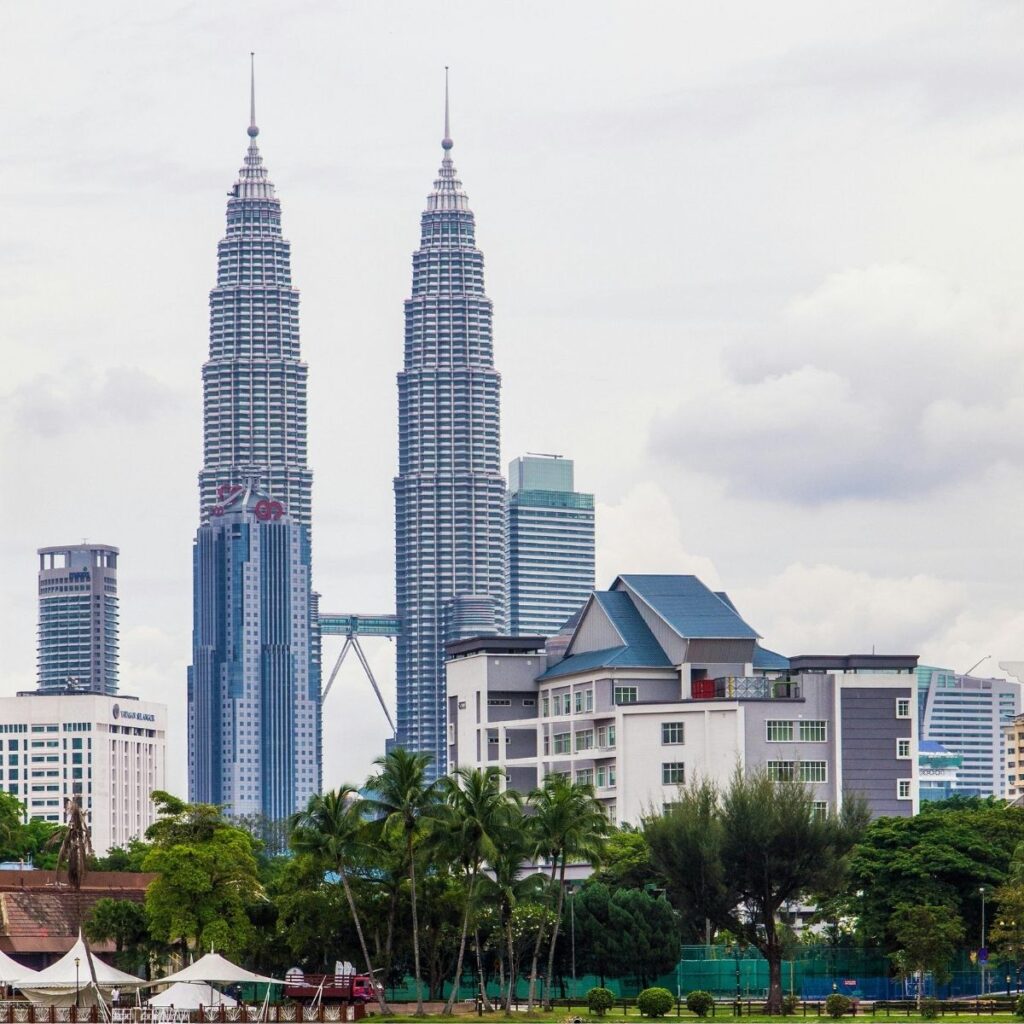 A low-angle view of the iconic Petronas Twin Towers in Kuala Lumpur, Malaysia, captured from across a scenic canal with traditional low-rise buildings in the foreground.