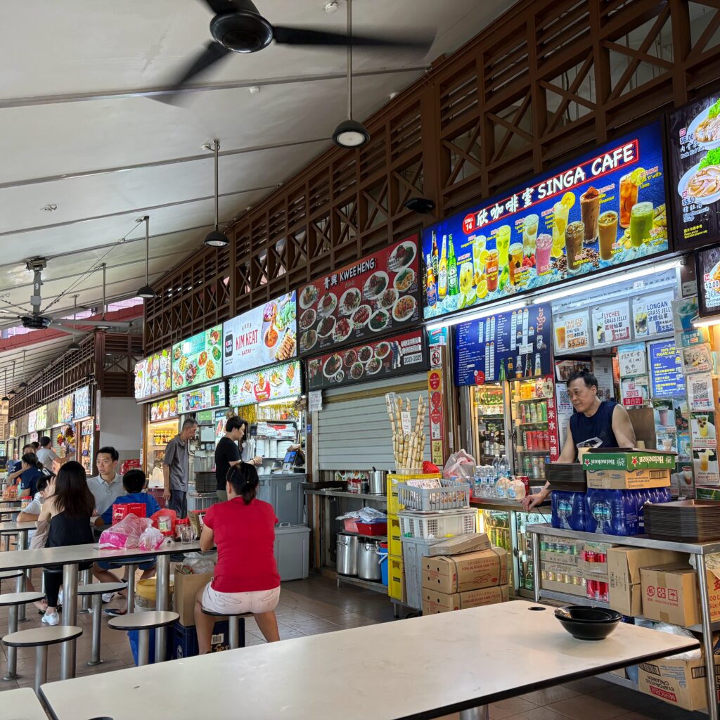 nterior view of a clean, bustling indoor Singapore hawker centre with rows of local food stalls and accessible dining tables.