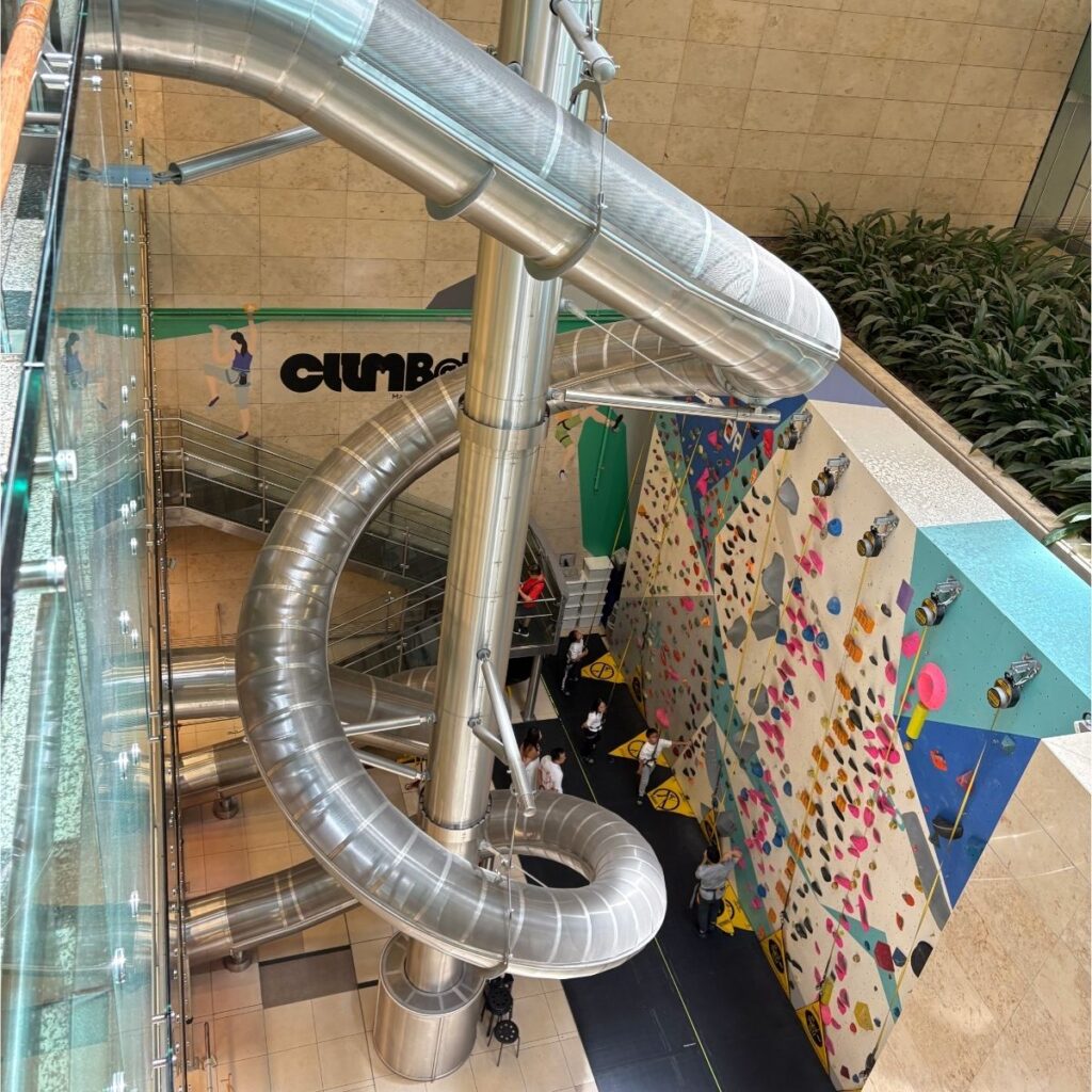 A wide multi-story indoor slide running next to a glass elevator inside a modern terminal at Singapore Changi Airport.