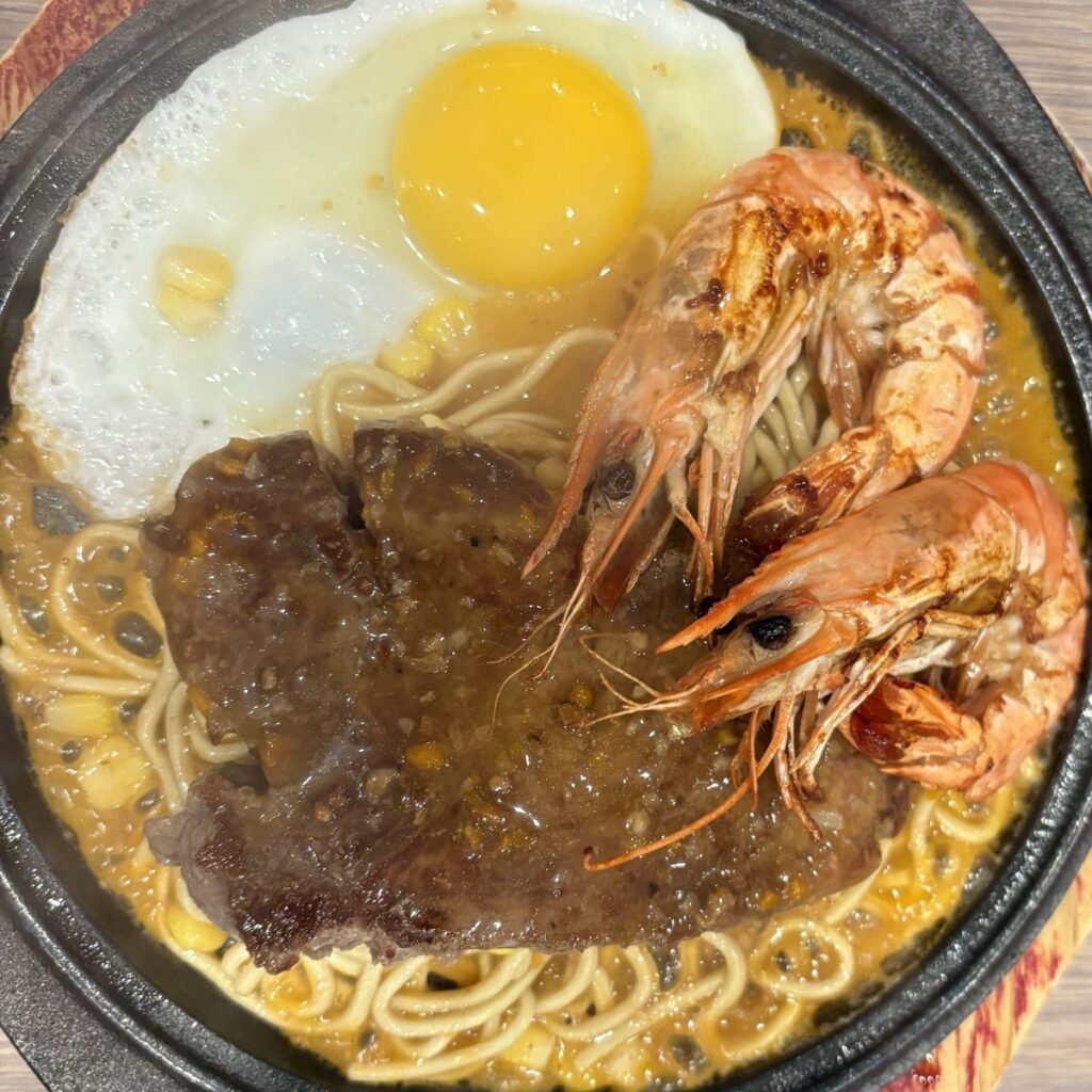 A classic Taiwanese-style steak served on a sizzling hot plate with noodles, two prawns, and a fresh egg.