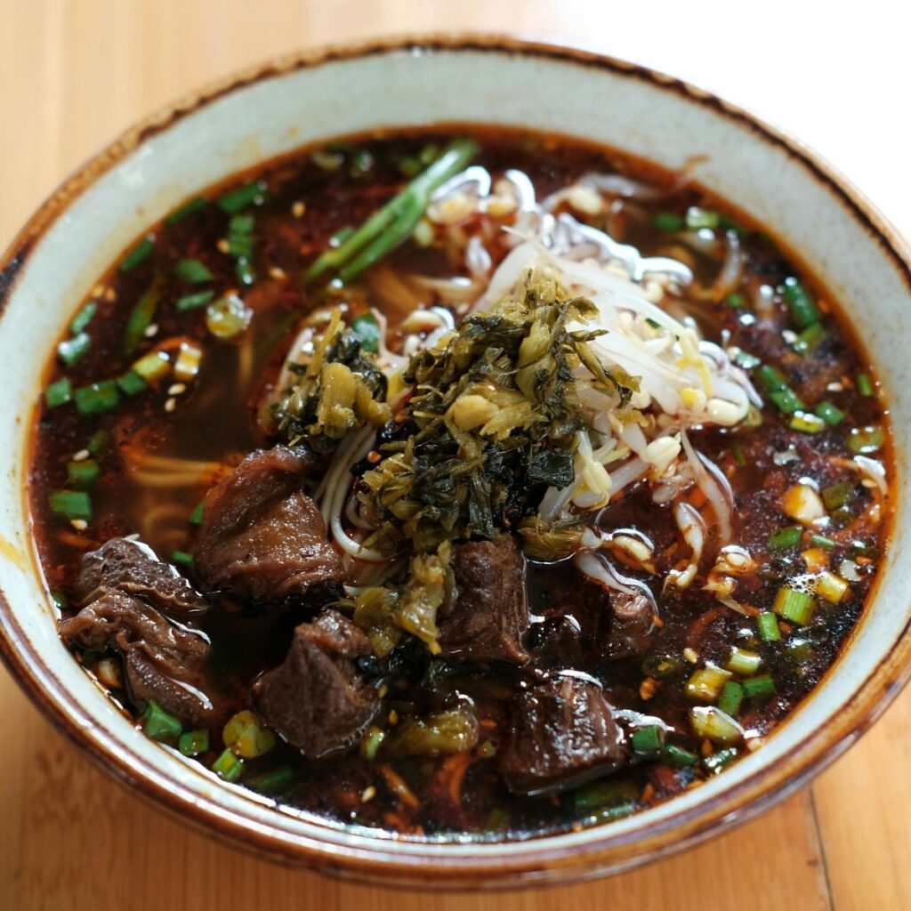 A steaming bowl of traditional Taiwanese beef noodle soup topped with fresh green onions and sour pickled vegetables.