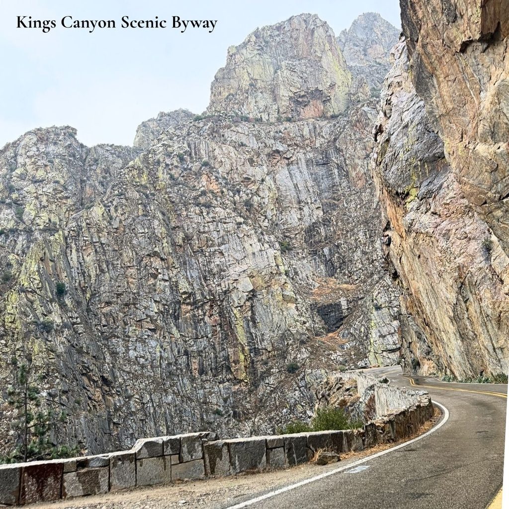 A winding curve on the Kings Canyon Scenic Byway flanked by towering, sheer granite mountain walls under a bright sky.