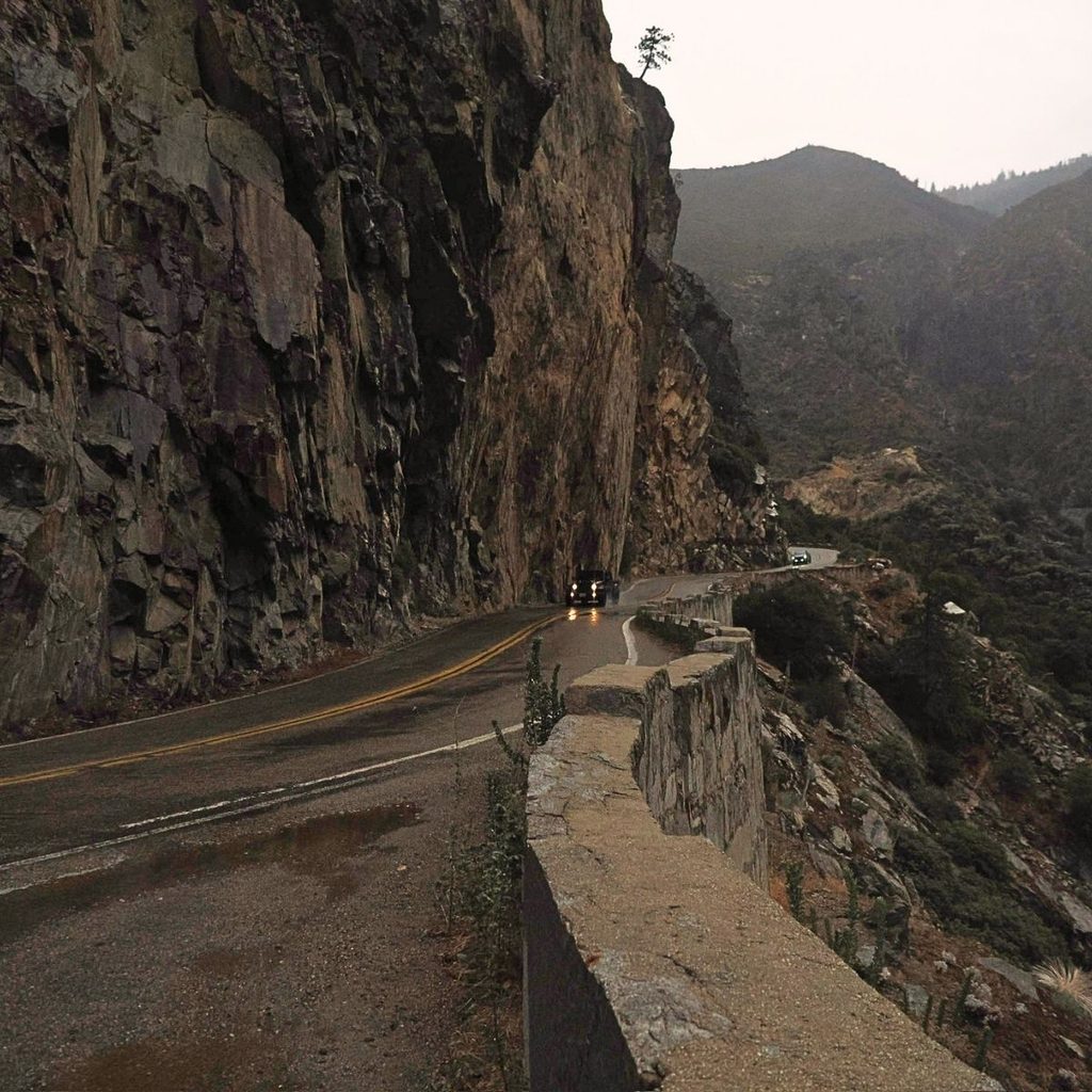 A dark, narrow mountain highway hugging a sheer vertical granite cliff wall with a stone guardrail along a steep valley ledge.