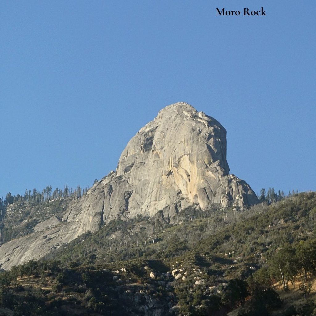 A majestic, sweeping low-angle view of the massive Moro Rock granite dome rising steeply against a clear blue sky in Sequoia National Park.