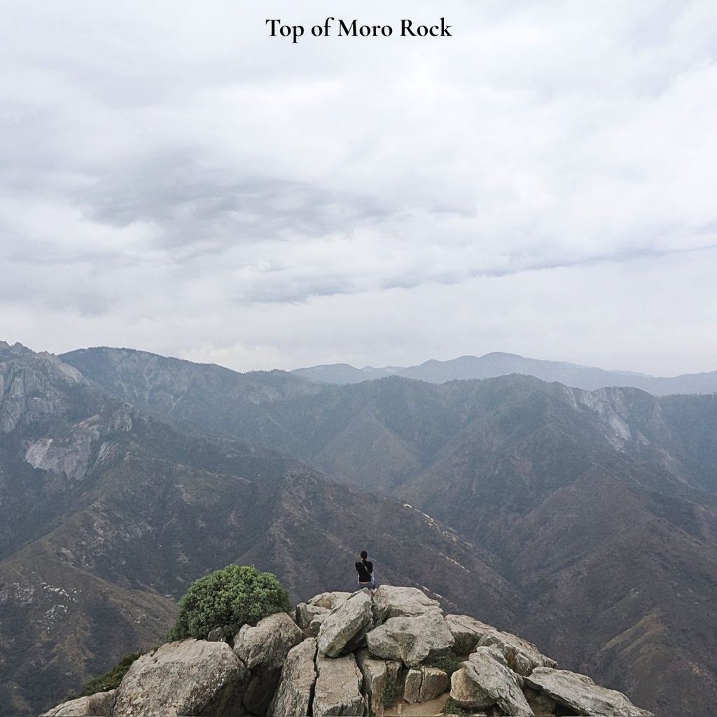 A person sitting peacefully on the rocky granite summit of Moro Rock looking out over the vast, expansive mountain ridges of Sequoia National Park under a cloudy sky.